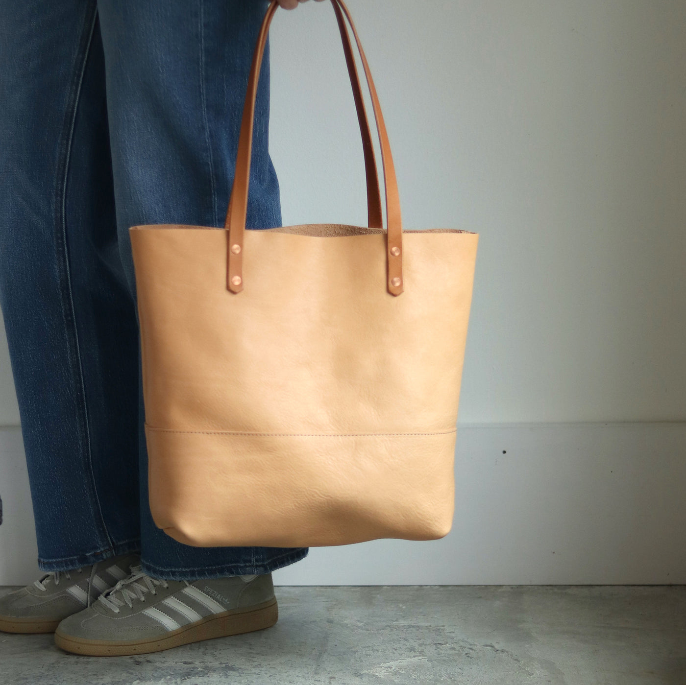 Beige leather tote held next to woman in casual blue jeans and Adidas sneakers