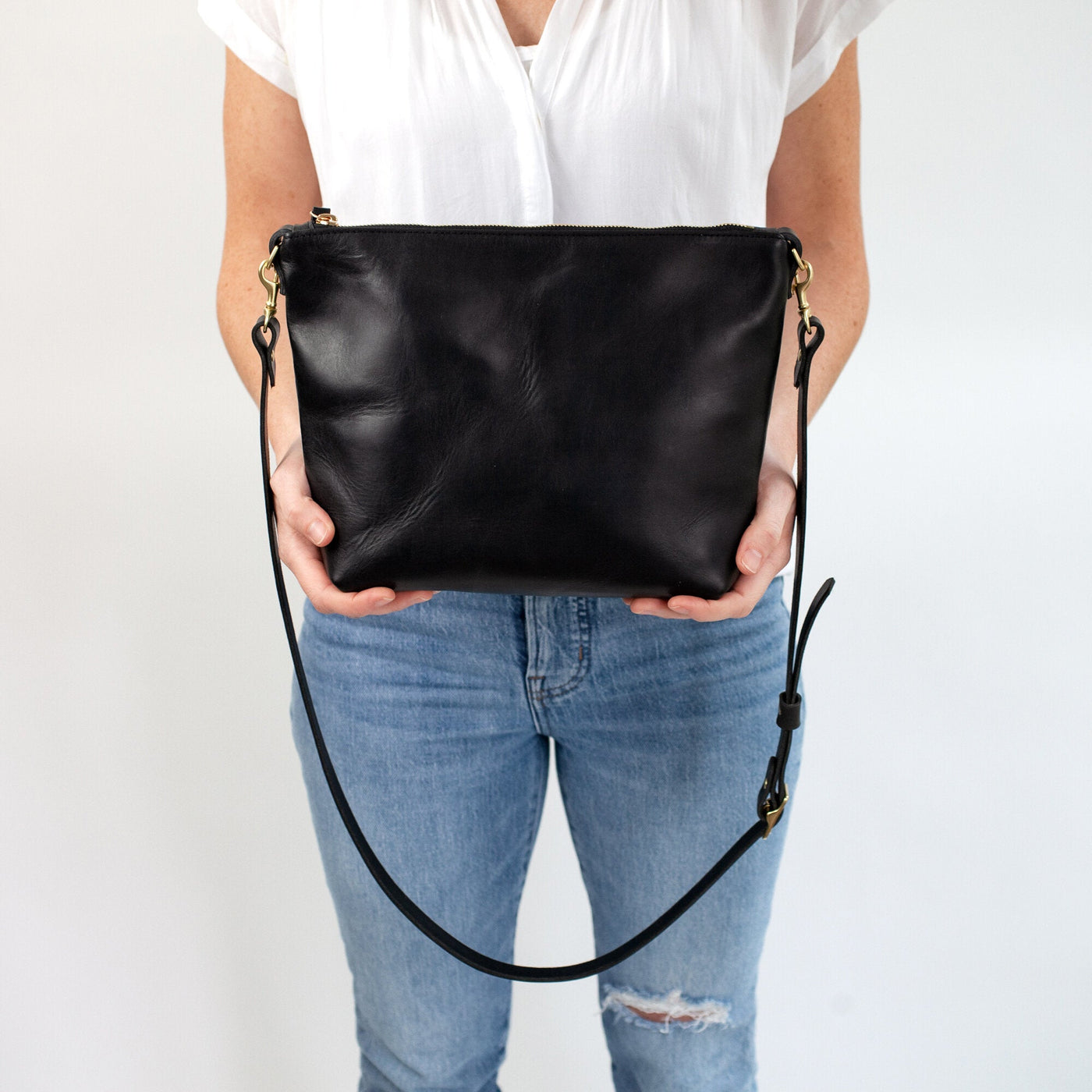 Woman in casual outfit of distressed jeans and white shirt holding a premium black leather handbag with top zipper closure and adjustable strap.  The bag is minimalistic and simple, and handmade with vegetable tanned leather.