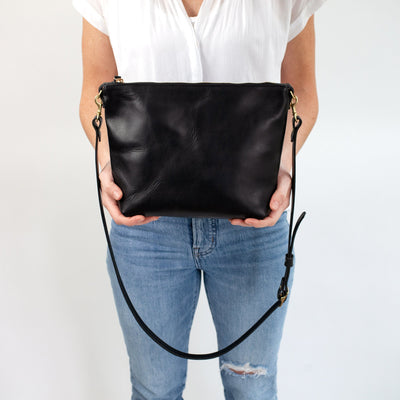 Woman in casual outfit of distressed jeans and white shirt holding a premium black leather handbag with top zipper closure and adjustable strap.  The bag is minimalistic and simple, and handmade with vegetable tanned leather.