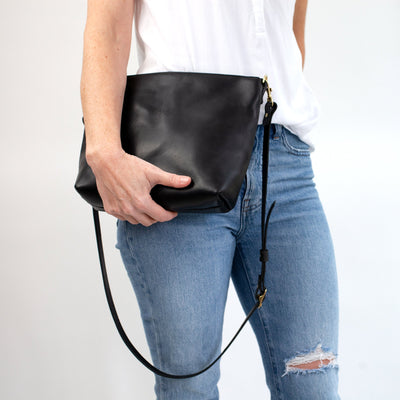 Women's minimalist black leather crossbody bag with top zipper and adjustable and removable strap. Converts to oversize clutch. Handmade with vegetable tanned leather and solid brass hardware. Woman in casual outfit of jeans and white shirt with casual black everyday leather handbag.