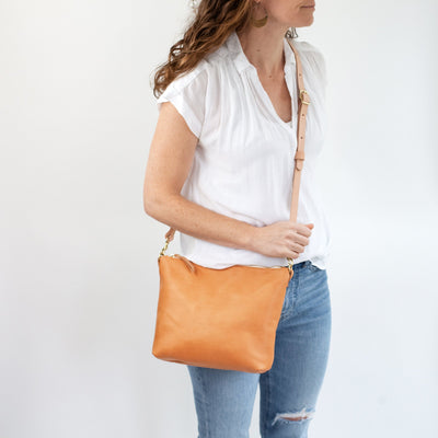 Women's beige color vegetable tanned leather bag, handmade with premium vegetable tanned leather and solid brass hardware. Top zipper closure, adjustable and removable strap, and available with a monogram.  Woman in casual outfit of jeans and white shirt with minimalist beige leather bag.